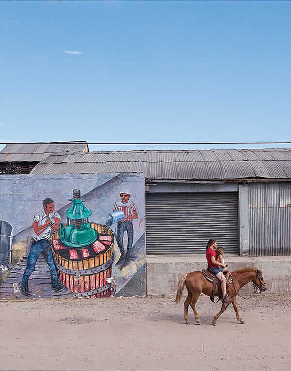 Two locals ride by La Escuelita, whose exterior is made of glass bottles, repurposed wine barrels and mattress springs.