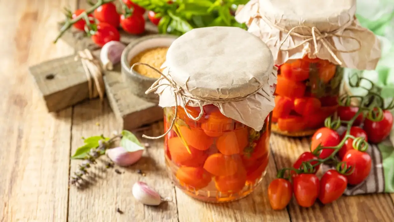 On a wooden table there are two jars of canned pickled cherry tomatoes, next to them lies a ripe cluster of small, round, bright red fruits, several cloves of garlic and various spices.