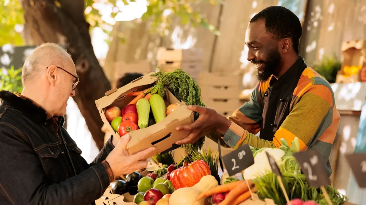 An elderly man buys various seasonal vegetables in a wooden box from a vendor at a farmers market.