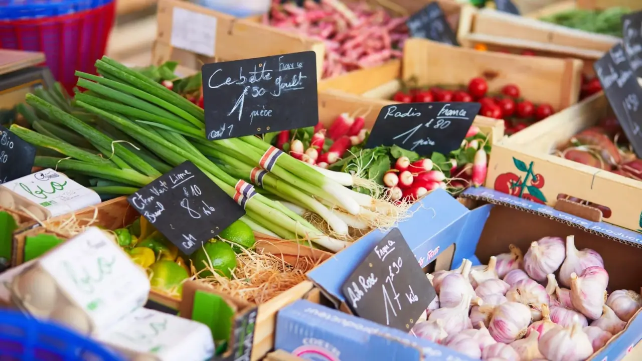Close up of market stall with cardboard boxes full of various vegetables with black signs stating product name and price.