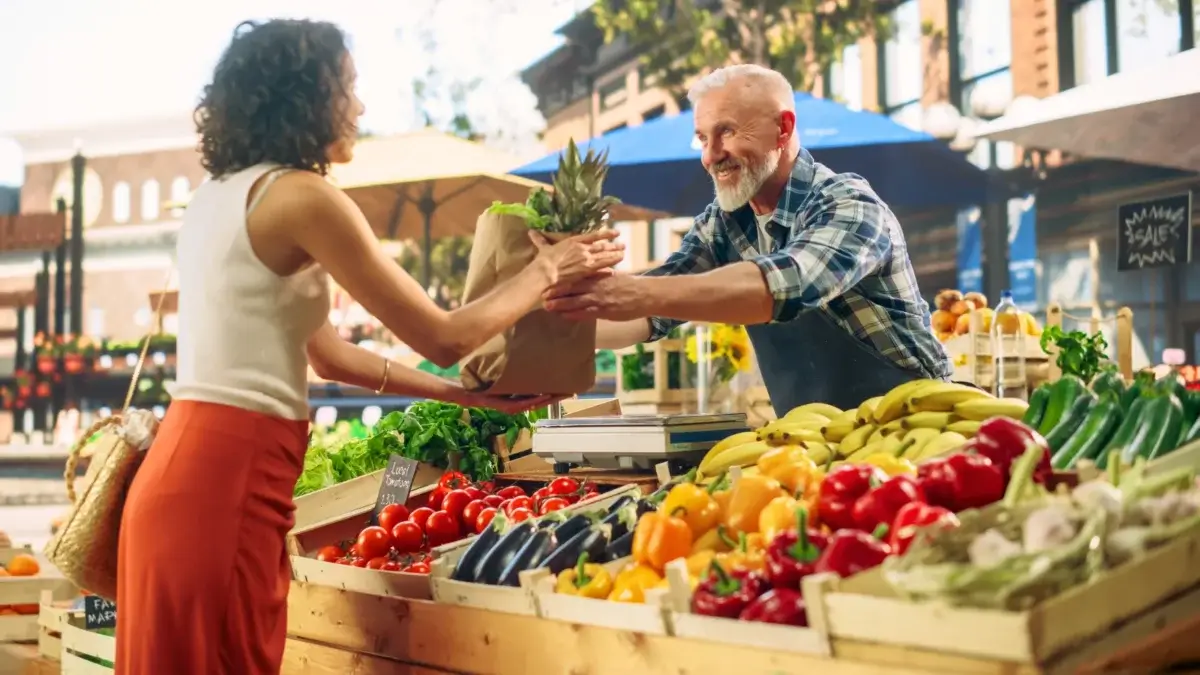 farmer's market tips. A farmer sells a paper bag full of vegetables and fruits to a young woman standing at a large counter with wooden boxes filled with various crops at a market.
