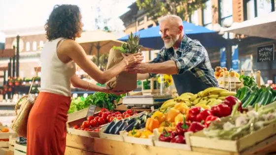 farmer's market tips. A farmer sells a paper bag full of vegetables and fruits to a young woman standing at a large counter with wooden boxes filled with various crops at a market.