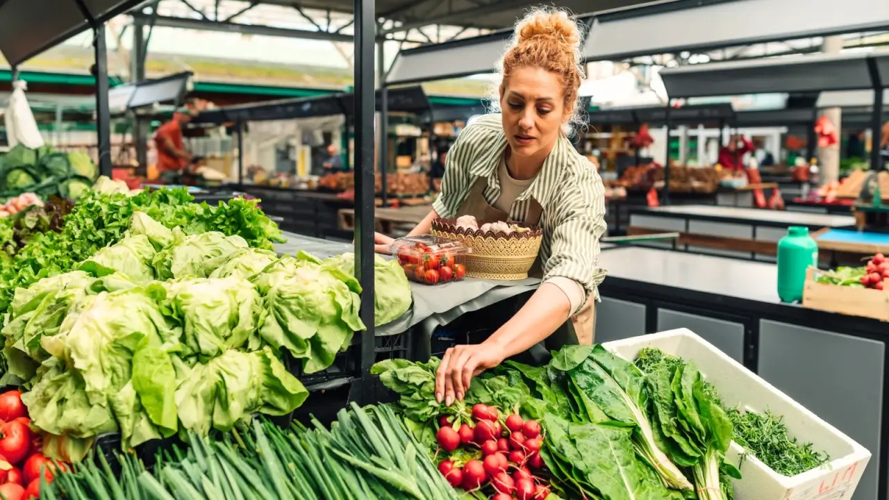 A female vendor in a striped apron checks her vegetables and greens laid out on a counter at a farmers market.