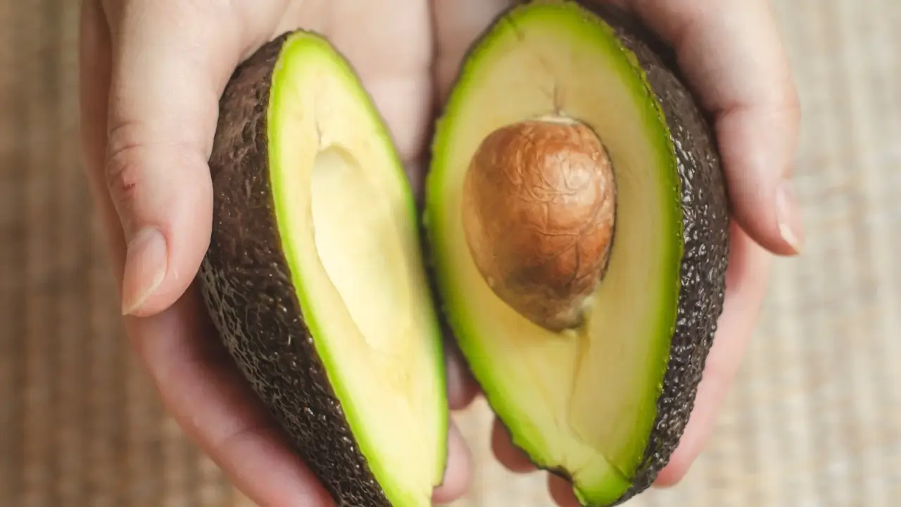 A hand holds a ripe avocado cut in half, showing its smooth green flesh and large brown seed in the center.
