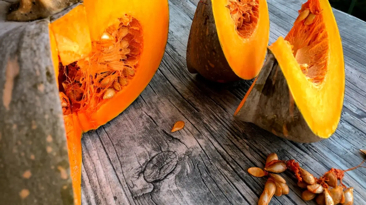 A pumpkin cut into pieces with orange flesh and seeds scattered on a wooden table.