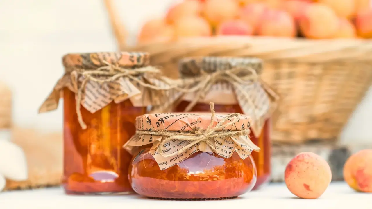 Glass jars of apricot jam stand against the backdrop of a large basket full of fresh apricots.