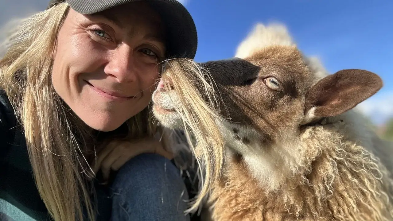 A photo of Melissa Schneider with an animal, appearing to sniff her hair while Melissa looks into the camera with the clear blue sky in the background