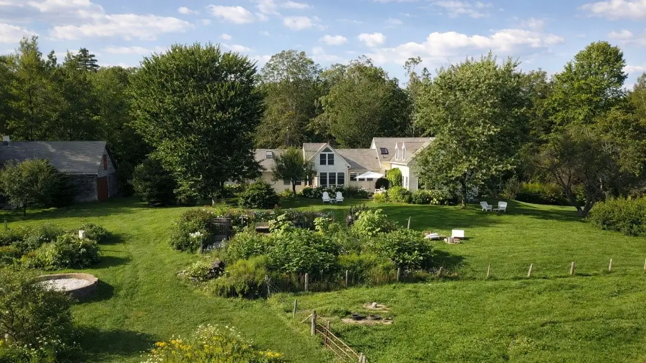 A wide shot of the property with a lovely house sitting among bright green grass and trees, looking lovely under bright and warm sunlight